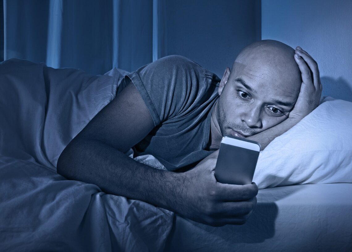 young cell phone addict man awake at night in bed using smartphone for chatting flirting and sending text message in internet addiction and mobile abuse concept