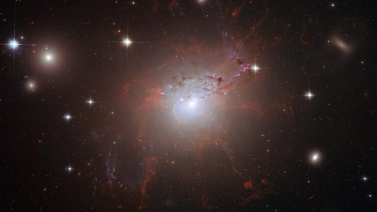 Galáxia NGC 1275 com núcleo brilhante, filamentos avermelhados difusos e estrelas em primeiro plano sobre o céu escuro