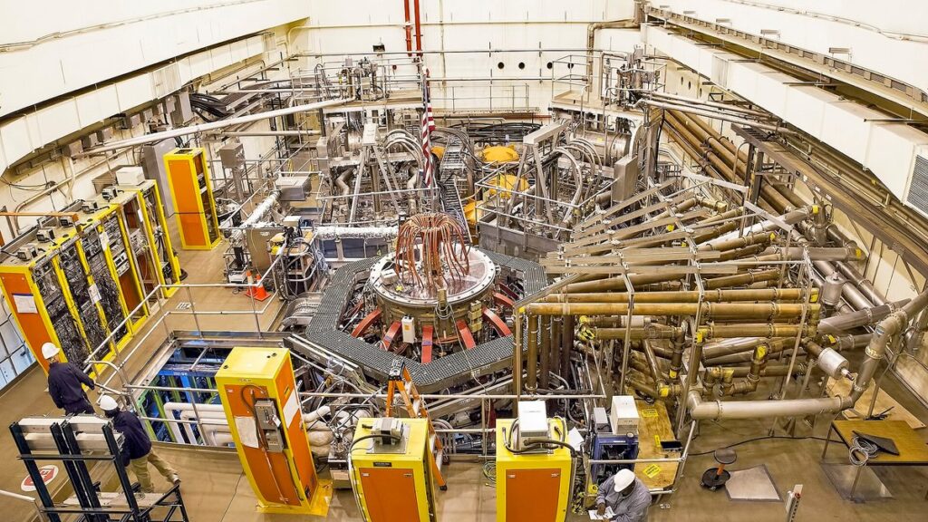 Vista superior do reator NSTX-U, com bobinas de cobre, tubulações metálicas, armários amarelos e técnicos de capacete.