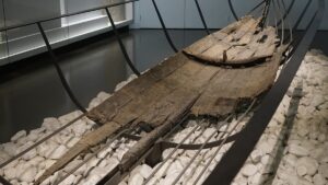 Casco de madeira escurecida do barco grego Jules-Verne 9, com tábuas unidas, apoiado sobre pedras brancas em exposição.