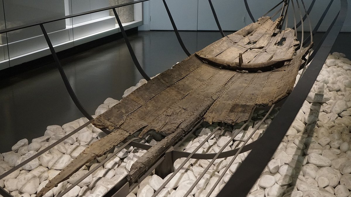 Casco de madeira escurecida do barco grego Jules-Verne 9, com tábuas unidas, apoiado sobre pedras brancas em exposição.