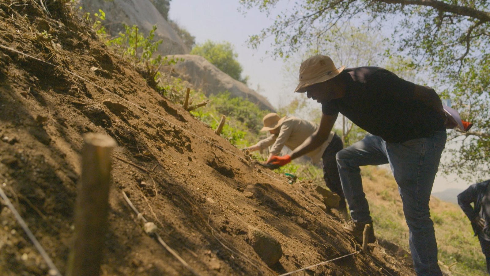 Dois homens de chapéu escavam com as mãos uma encosta de terra íngreme, com cordas de demarcação e vegetação ao fundo.