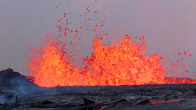Fonte de lava laranja e incandescente jorrando alto sobre uma superfície escura de rocha vulcânica sob céu acinzentado.