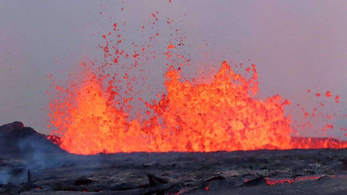 Fonte de lava laranja e incandescente jorrando alto sobre uma superfície escura de rocha vulcânica sob céu acinzentado.