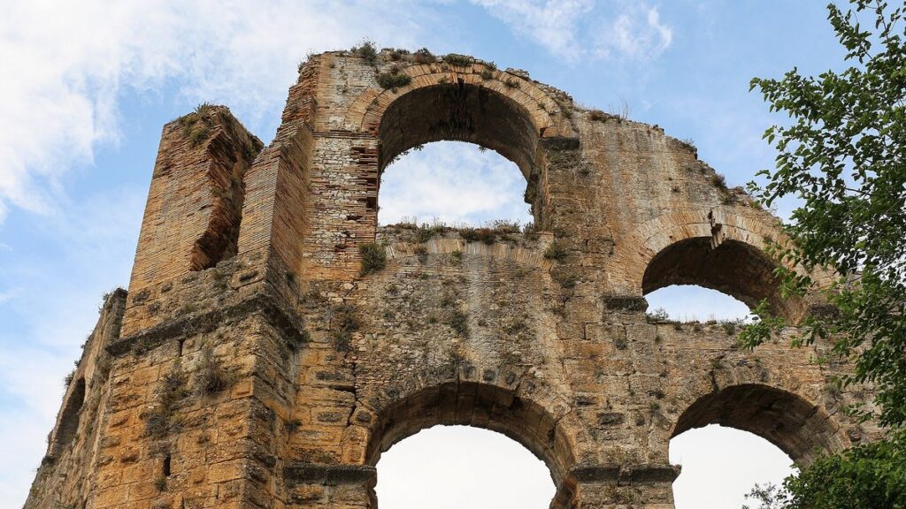 Ruína de aqueduto com arcos de pedra e tijolo sob céu azul, vista de baixo para cima, com folhagem à direita.