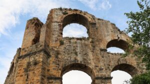 Ruína de aqueduto com arcos de pedra e tijolo sob céu azul, vista de baixo para cima, com folhagem à direita.