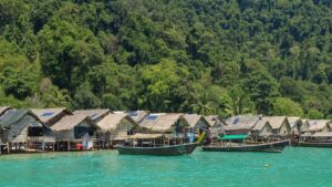 Casas de telhado de palha sobre palafitas na água turquesa, com barcos ancorados e encosta coberta de mata