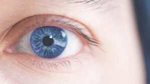 Close-up de um olho de íris azul, com reflexos de luz e cílios visíveis, sobre pele clara.