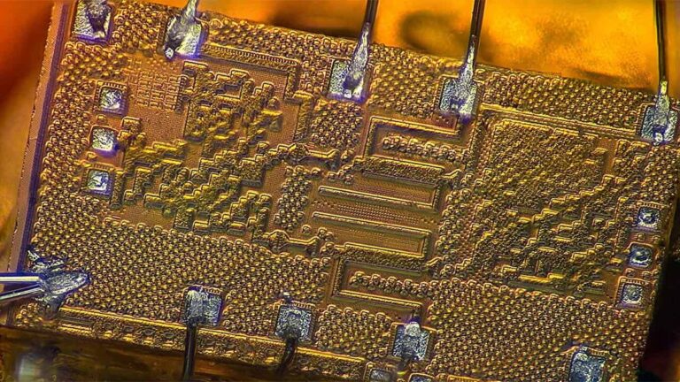 Microfotografia de um chip dourado com circuitos em relevo, contatos metálicos e fios finos ligados às bordas.