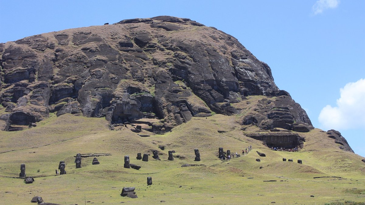 Estátuas moai de pedra espalhadas por uma encosta gramada, diante de um paredão rochoso sob céu azul.