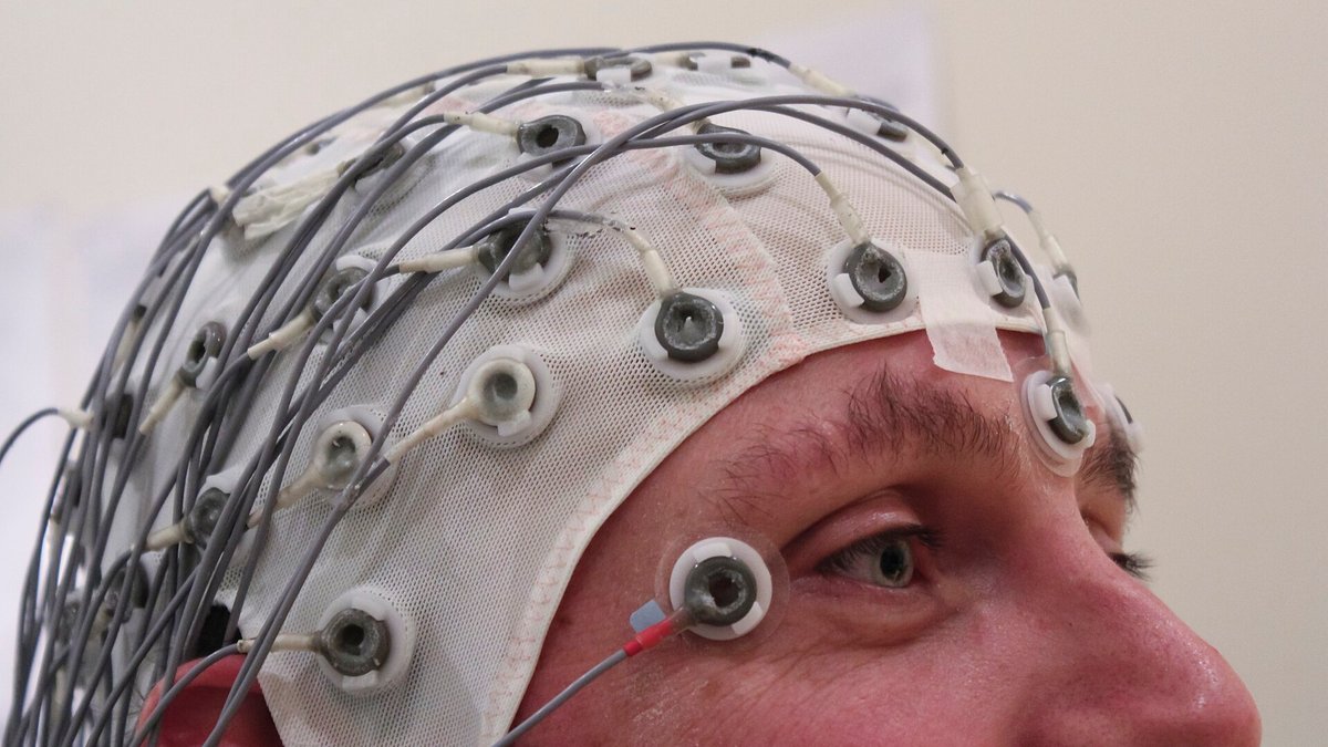 Perfil de uma pessoa com gorro branco de eletrodos e fios cinzas para registro de atividade cerebral por EEG.