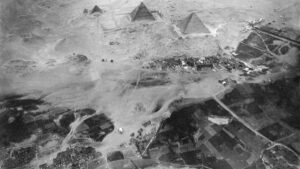 Foto aérea em preto e branco de três pirâmides no deserto, com povoado e campos ao redor