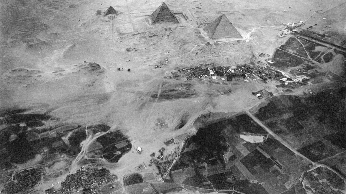 Foto aérea em preto e branco de três pirâmides no deserto, com povoado e campos ao redor