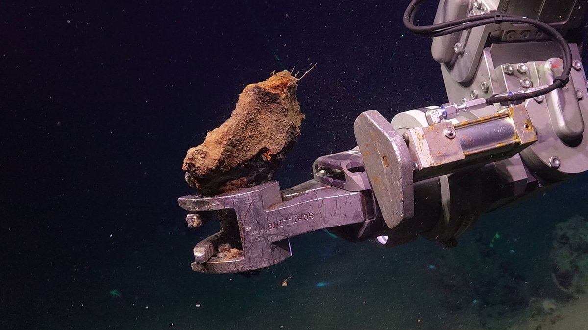 Braço robótico metálico submarino segura uma rocha áspera e enferrujada, com fundo escuro de água do mar ao redor.