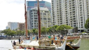 Canoa de dois cascos Hōkūleʻa atracada em porto, com pessoas no convés e prédios altos ao fundo.