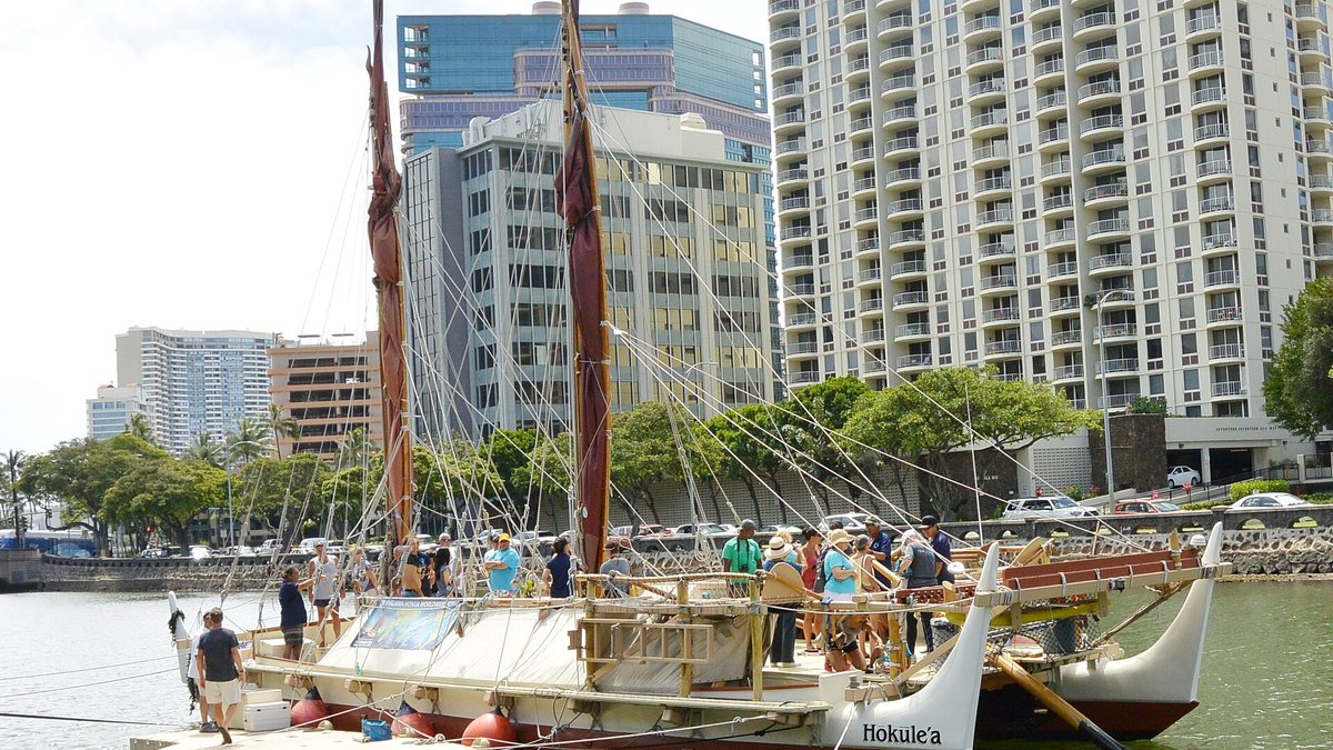Canoa de dois cascos Hōkūleʻa atracada em porto, com pessoas no convés e prédios altos ao fundo.