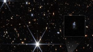 Campo de galáxias com estrelas brilhantes; no detalhe ampliado, pequena galáxia azulada marcada por cruz branca.