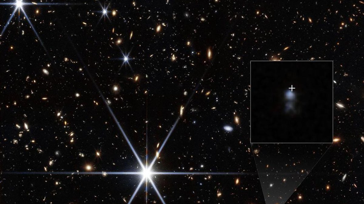 Campo de galáxias com estrelas brilhantes; no detalhe ampliado, pequena galáxia azulada marcada por cruz branca.